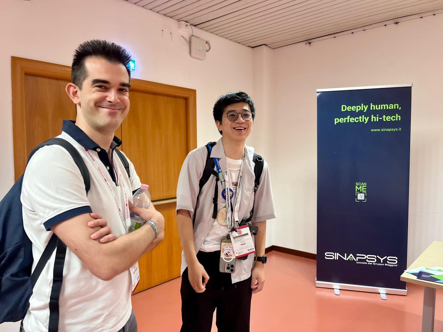 Due giovani partecipanti alla conferenza IEEE ICHI 2025 davanti allo stand Sinapsys, società del Gruppo Maggioli. Sullo sfondo, il roll-up aziendale con il claim “Deeply human, perfectly hi-tech”.