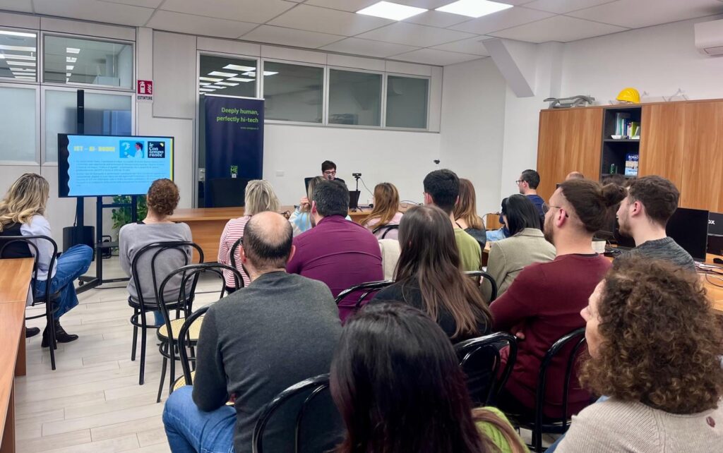 Evento formativo sulla parità di genere in un ambiente aziendale moderno. Un gruppo di persone ascolta un relatore che presenta una slide su uno schermo. L'aula ha un design professionale con sedie nere, scaffali in legno e un banner con il testo 'Deeply human, perfectly hi-tech'. Il pubblico è composto da uomini e donne attenti alla presentazione