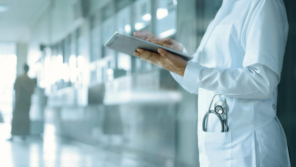 Dottoressa con camicie bianco in una corsia di un ospedale con in mano un tablet e in tasca uno stetoscopio