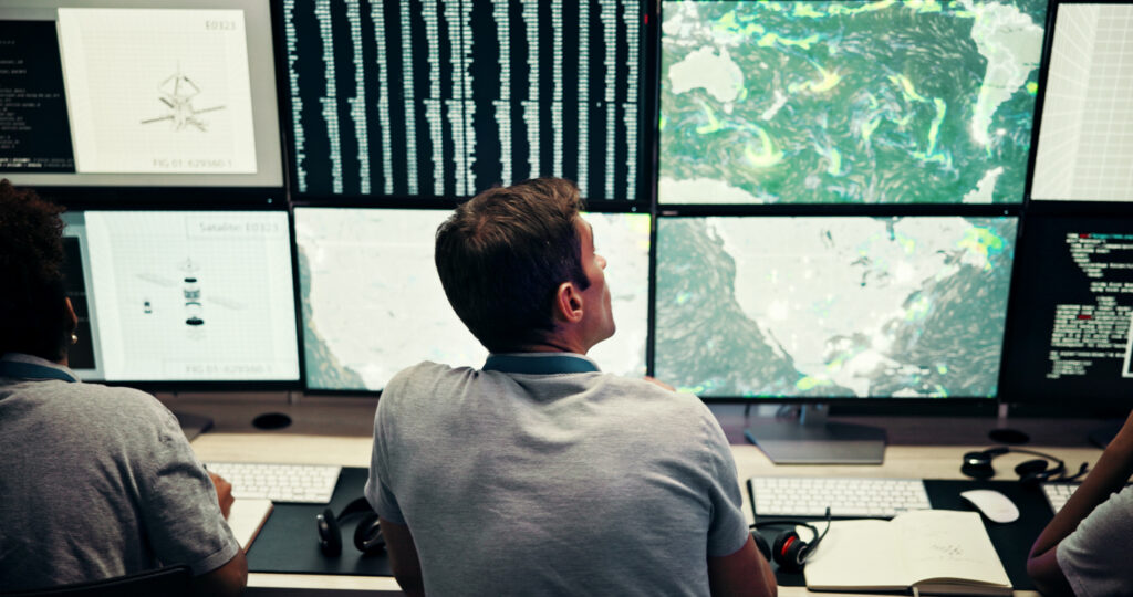 Operatori in una sala di monitoraggio osservano dati ambientali e mappe meteorologiche su più schermi per l’analisi e la previsione dei fenomeni climatici.