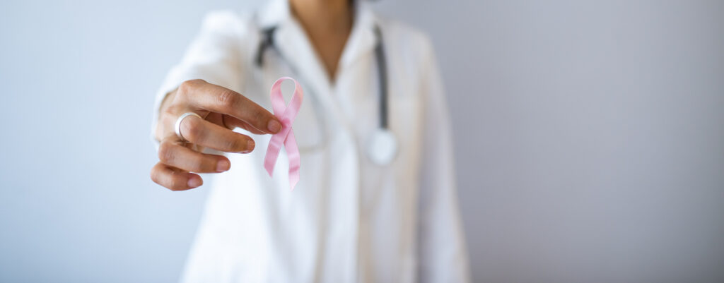 Una persona in camice bianco con stetoscopio mostra un nastro rosa, simbolo della sensibilizzazione sul tumore al seno.